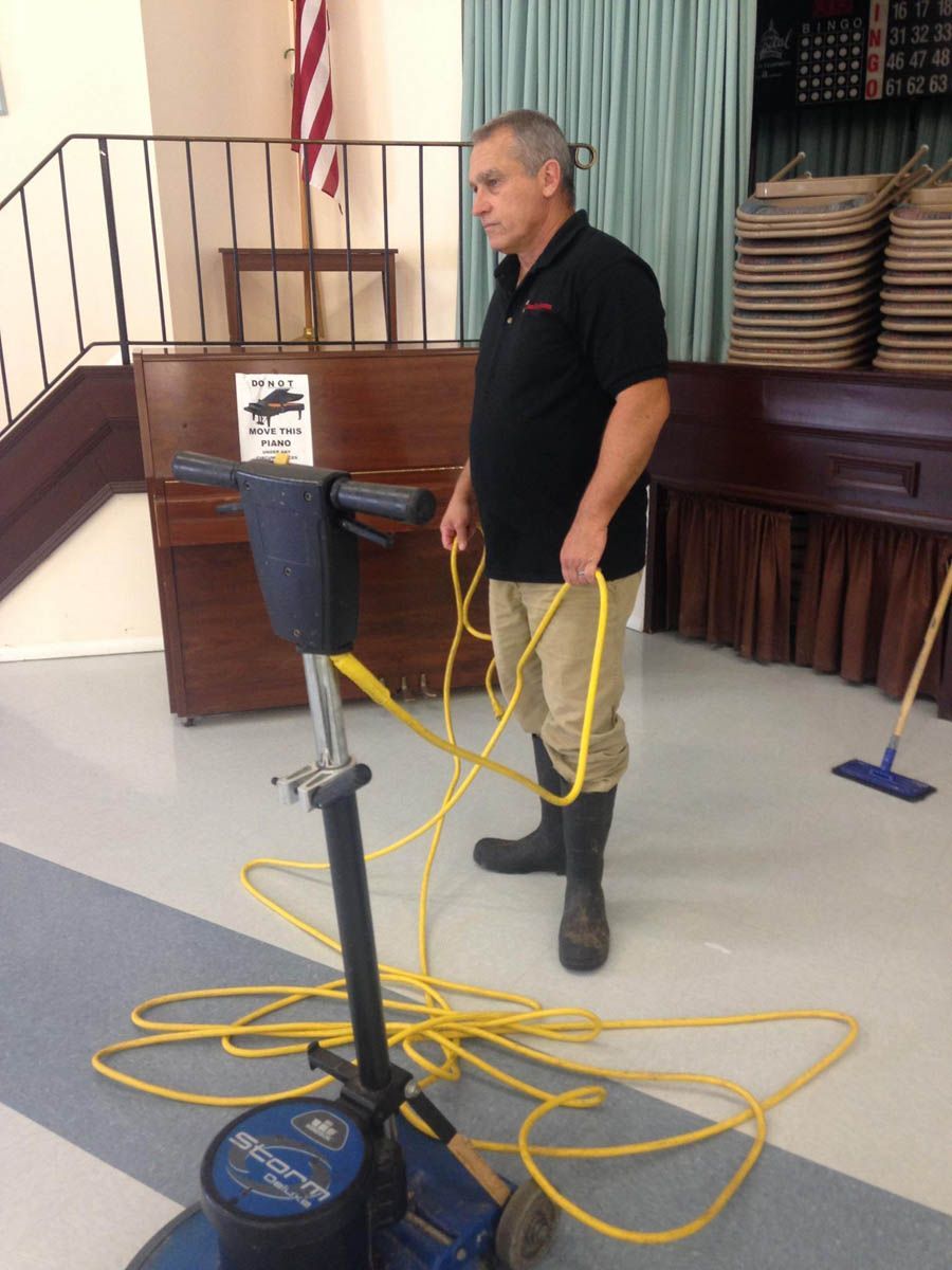 Man in black shirt and boots prepares floor buffer in a room with a piano and American flag.