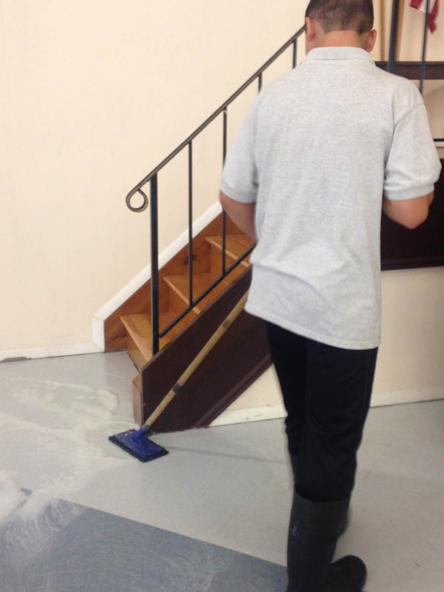 Person mopping a floor near a staircase. The floor is light grey. The stairs are wooden with a black railing.