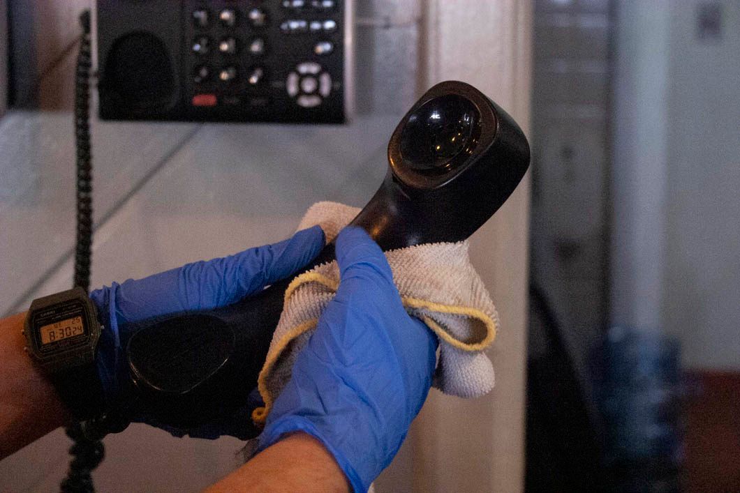 Hands in blue gloves wipe a black telephone receiver with a cloth near a wall-mounted keypad.