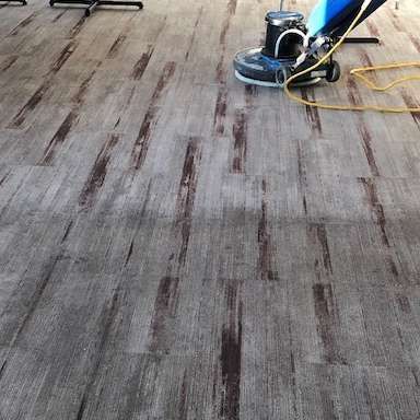A floor being cleaned with a machine. Wooden gray flooring with brown accents. The machine is blue and black.