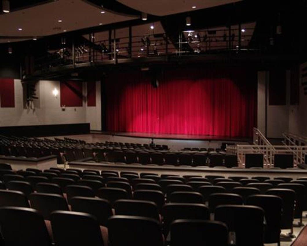 Auditorium interior with rows of seats facing a red stage curtain. Dark ceiling and walls, sound panels visible.