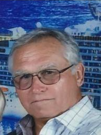 Man with glasses, wearing a light-colored plaid shirt, looks directly at the viewer. Background includes a ship.