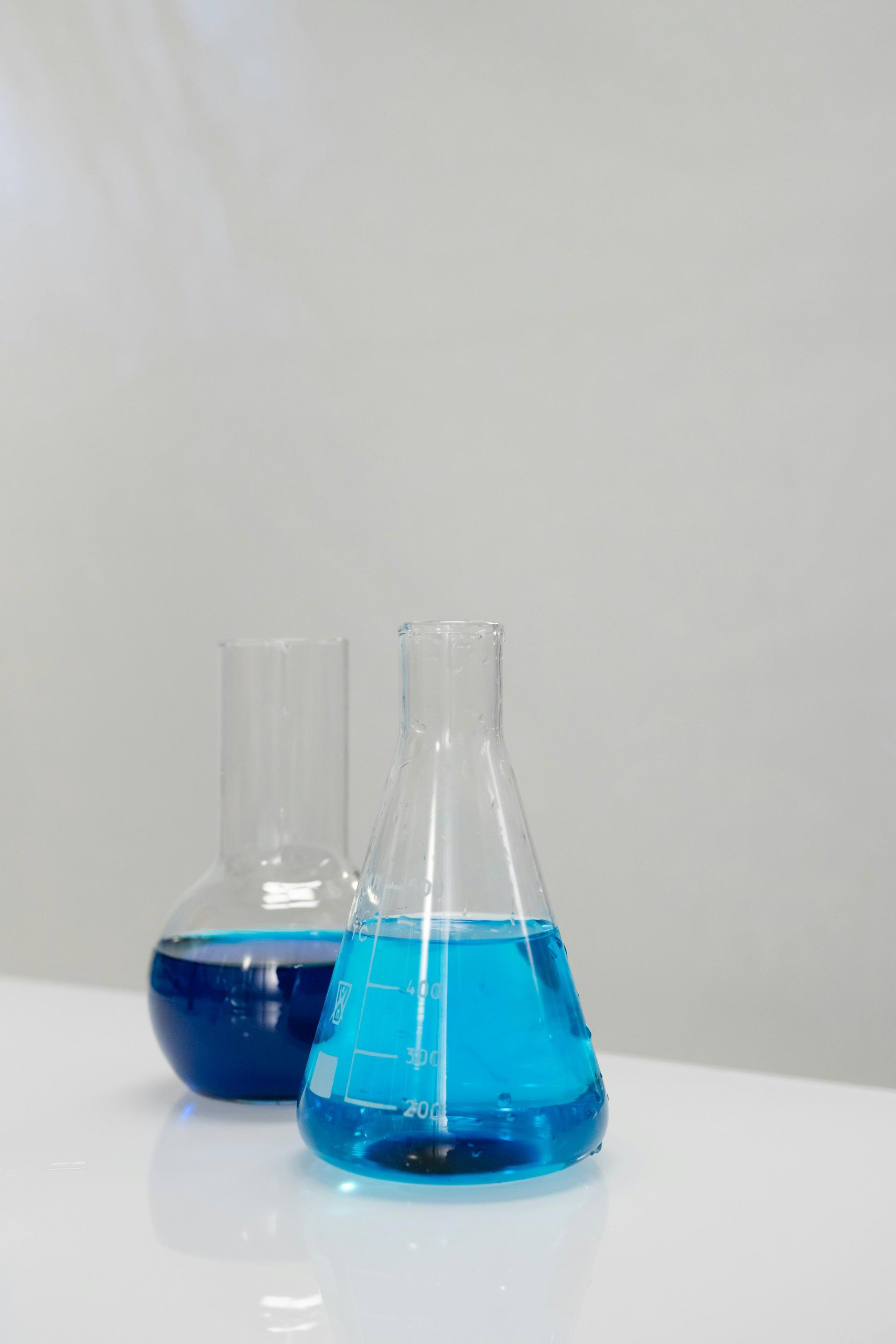 Two glass beakers with blue liquid, on a white surface, against a neutral background.