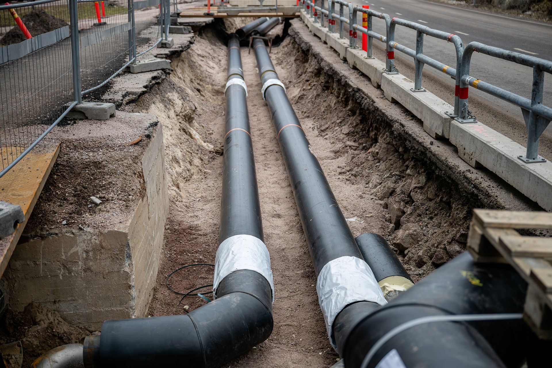 Trench with large insulated pipes under construction beside a road with safety railings