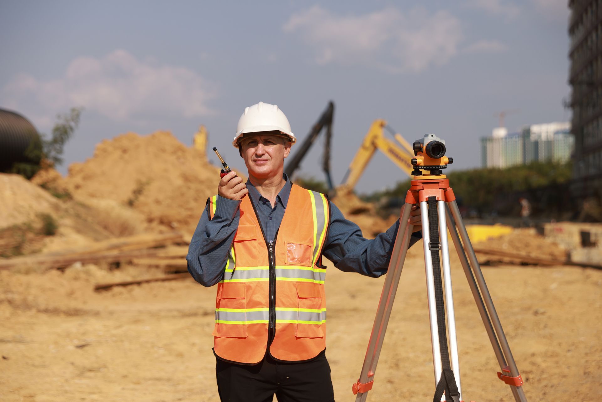 Surveyor in orange safety vest and hard hat beside tripod at a construction site, giving a thumbs-up.