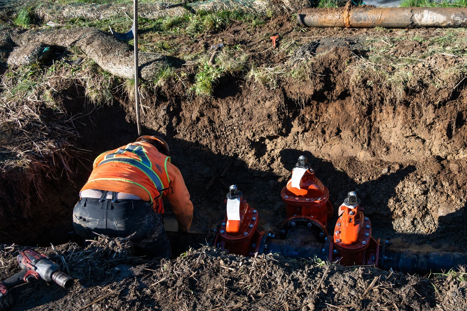 Workers in orange vests and hard hats inspect a trench beside a large pipe.