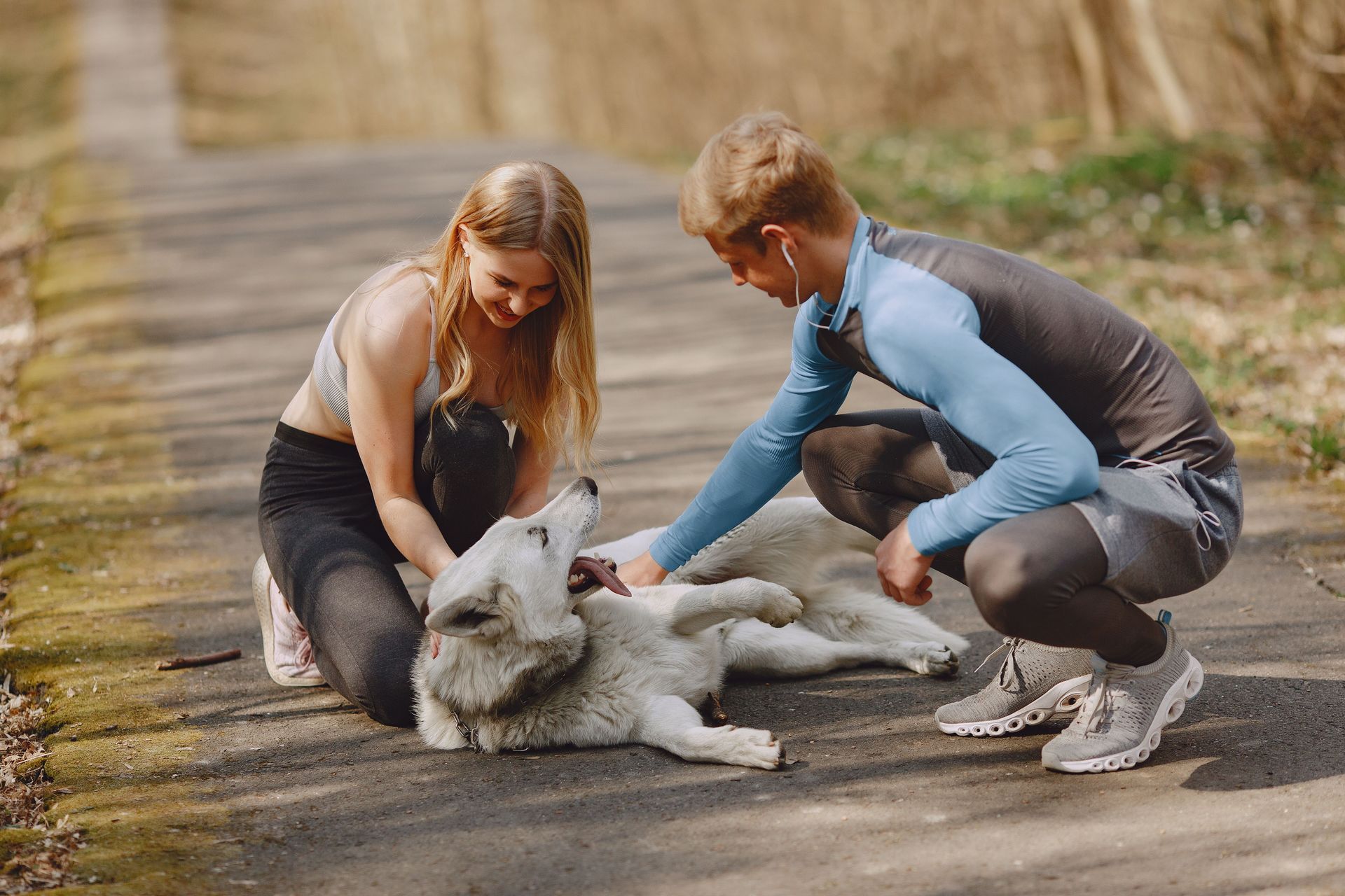 Two people pet a white dog outdoors on a rocky path in a wooded area