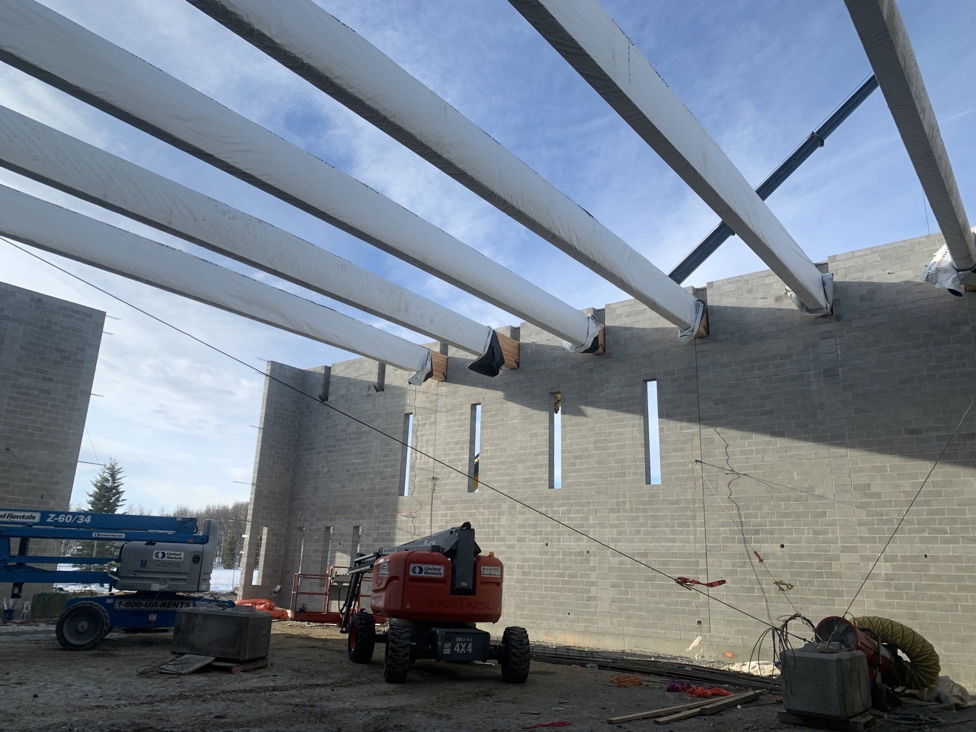 Steel beams being installed on a concrete building frame, with a lift truck below and blue sky overhead.