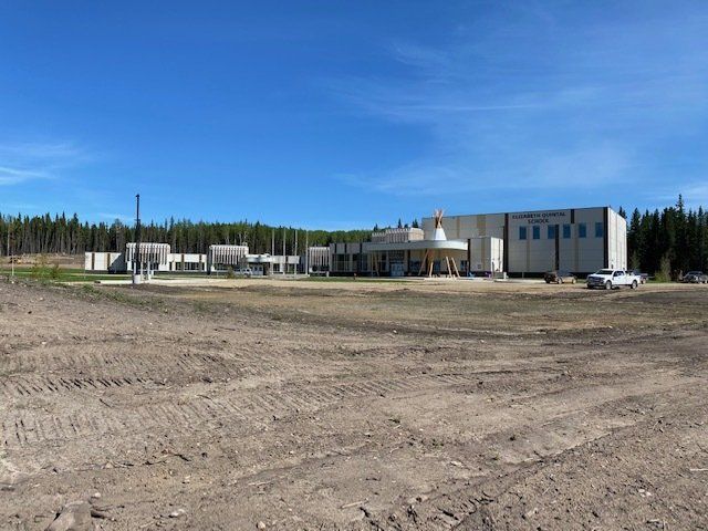 Large industrial building on a dirt lot under a blue sky, with trees in the background.