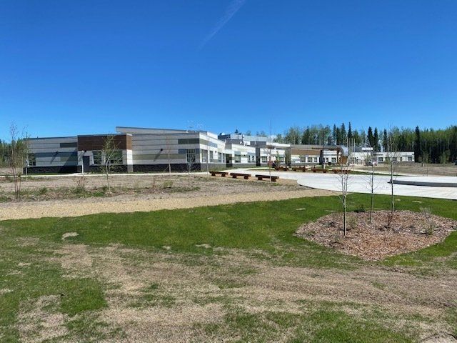 Modern low-rise building with a paved walkway, grassy foreground, and young trees under a clear blue sky