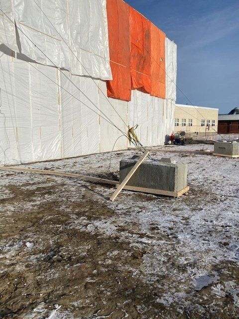 Snowy construction site beside white building wrapped in orange and white sheeting, with debris in foreground
