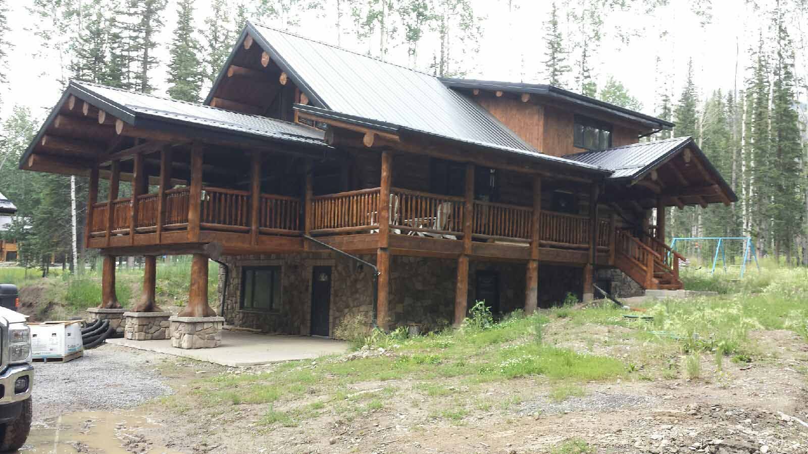 Wooden cabin with a large porch in a forest clearing, surrounded by tall trees and gravel ground