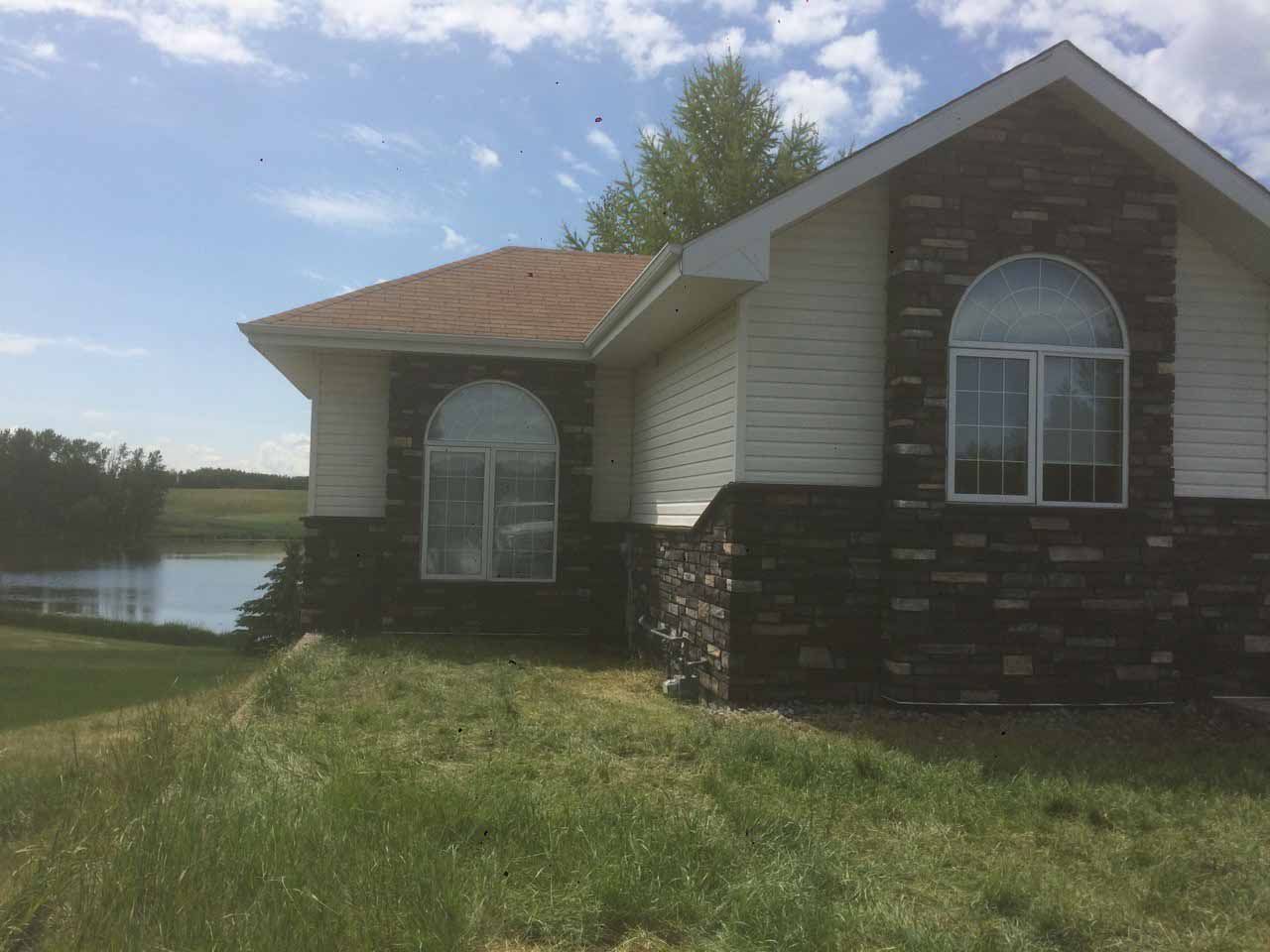 Rural house with brick and white siding beside a grassy yard and pond under a blue sky