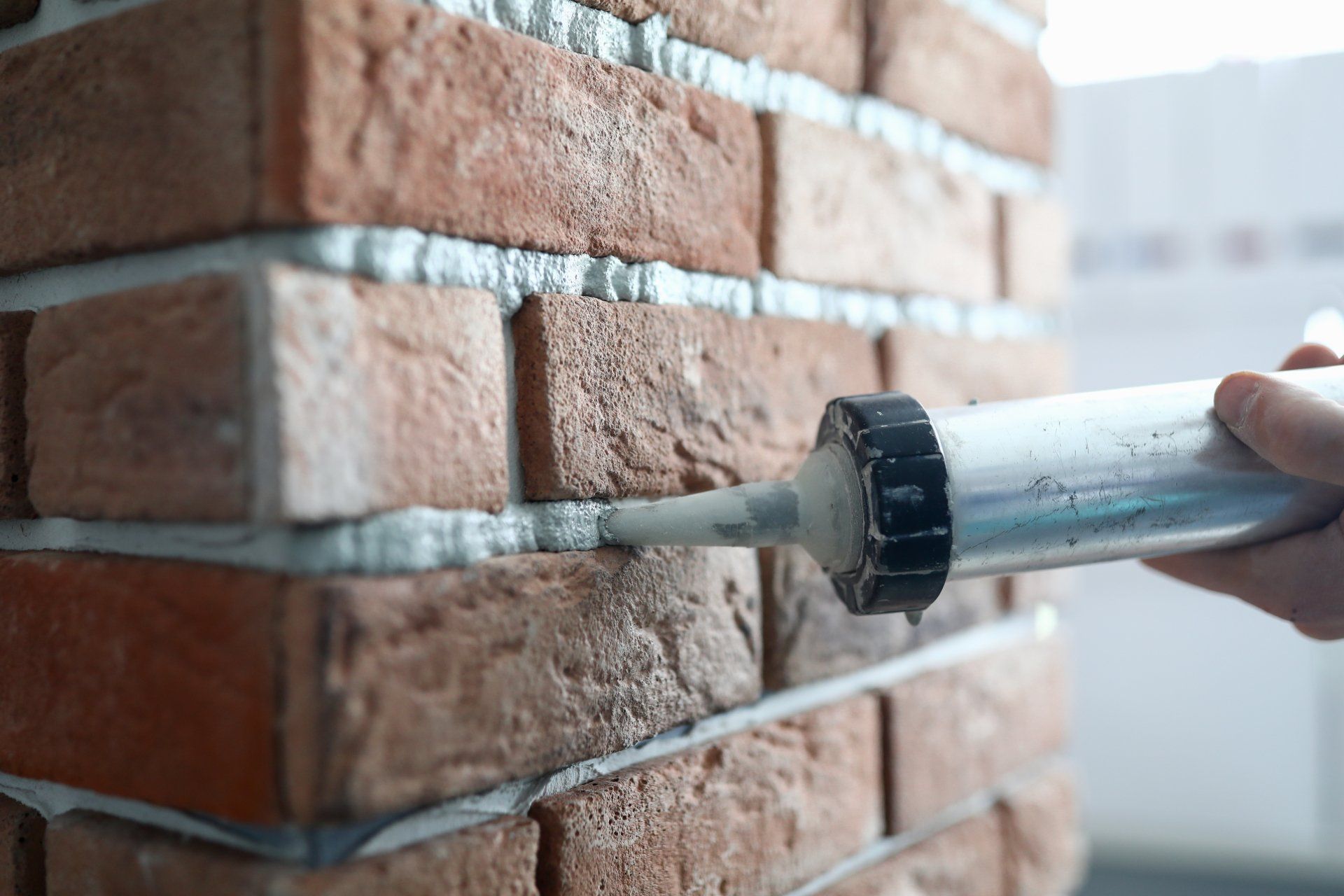 Hand holding a caulking gun applying sealant between bricks on a brick wall