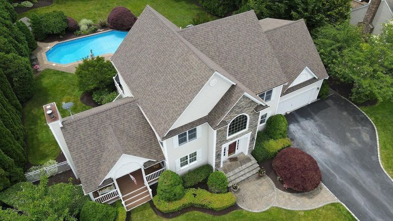 Aerial view of a large white house with a yard, driveway, patio, and swimming pool surrounded by trees.