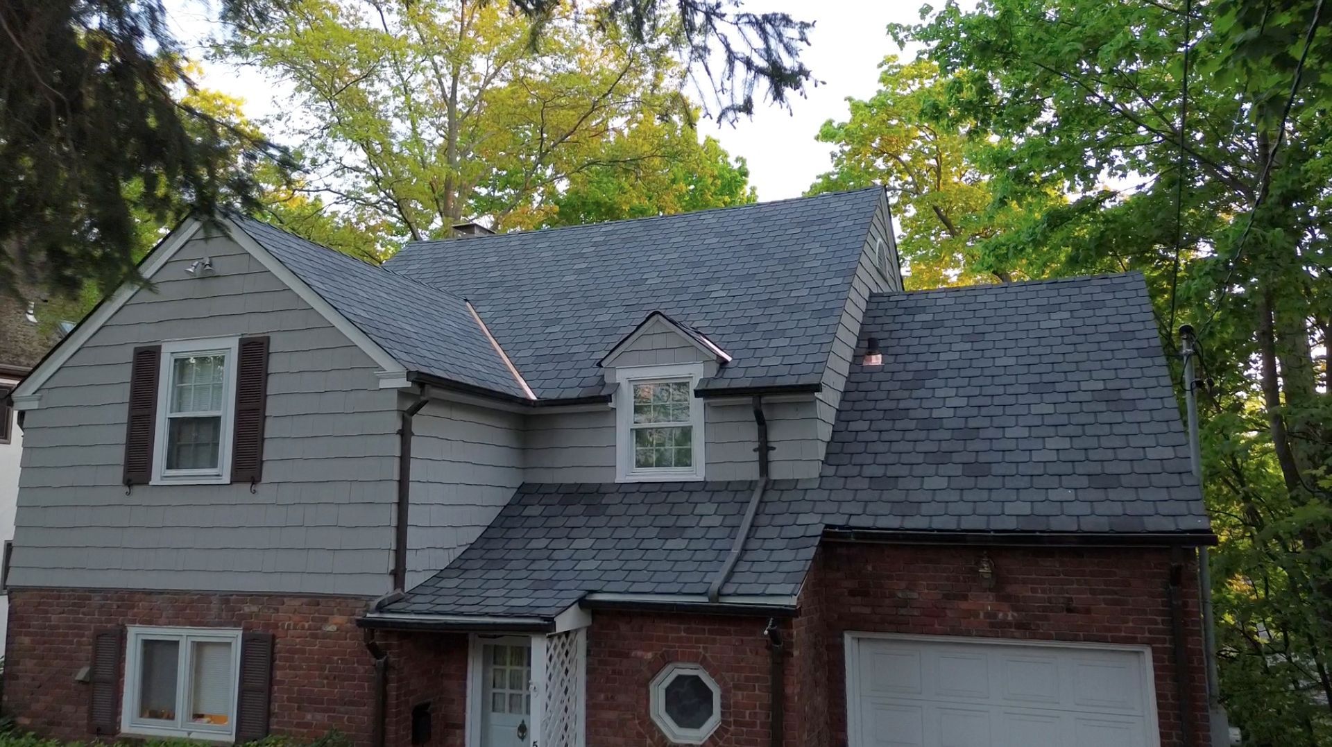Two-story house with gray siding, brick facade, dark roof, and attached garage surrounded by trees