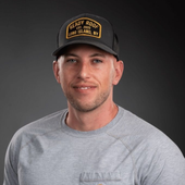 Man in gray shirt and black cap smiling against a dark studio background