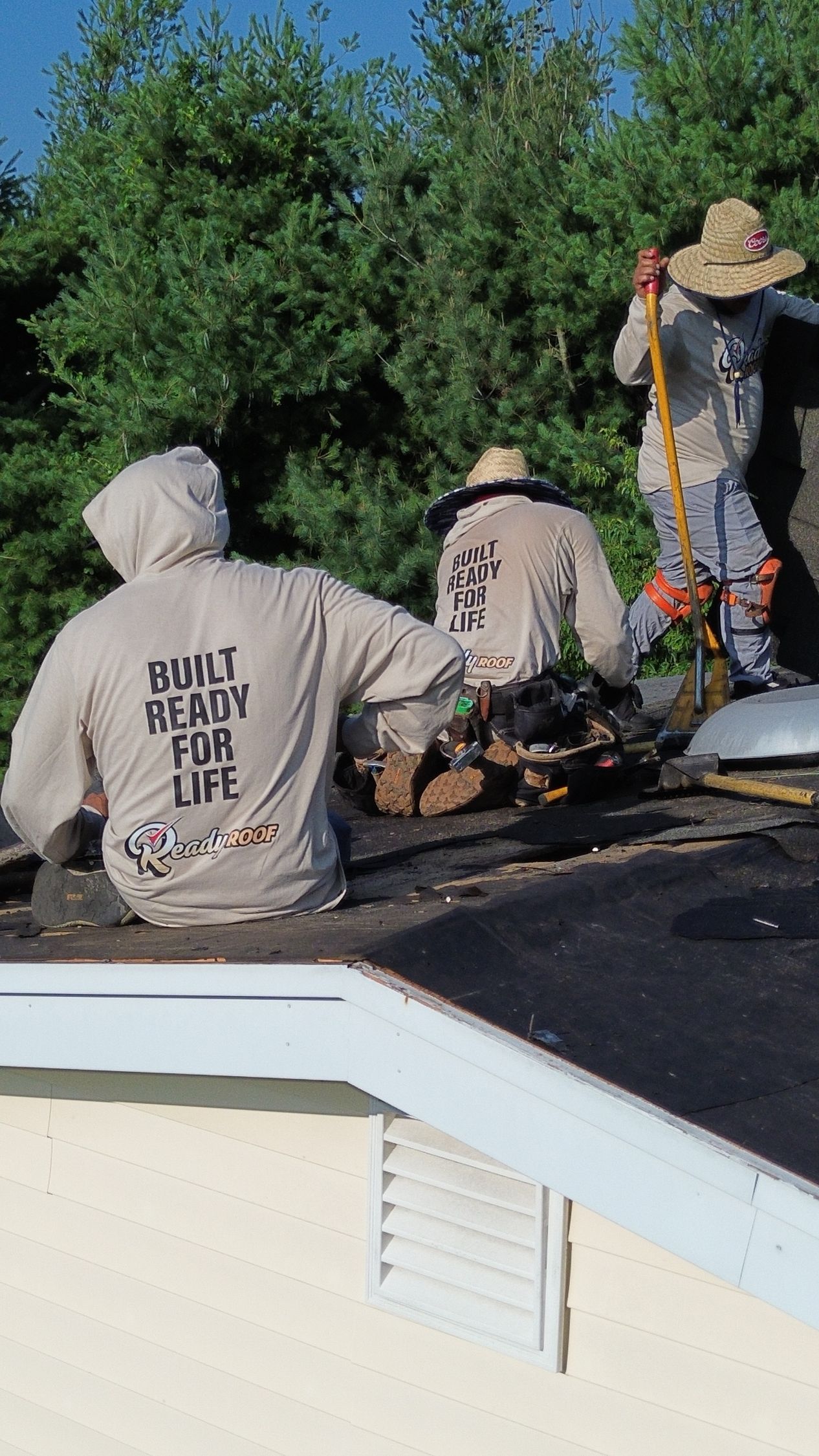 Workers repairing a roof beside a white house, with a man in a straw hat standing near tools and greenery.