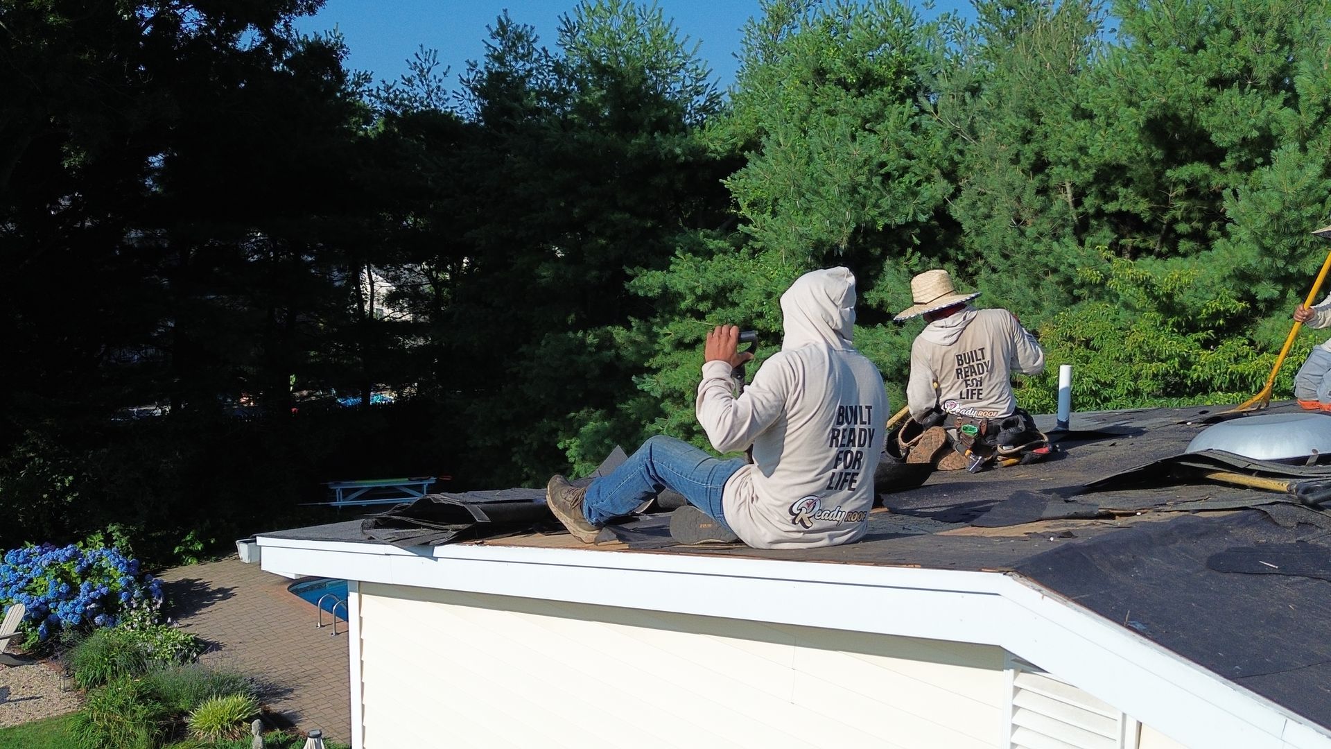 Two people sit on a rooftop near trees, one holding an orange drink, under a clear blue sky.