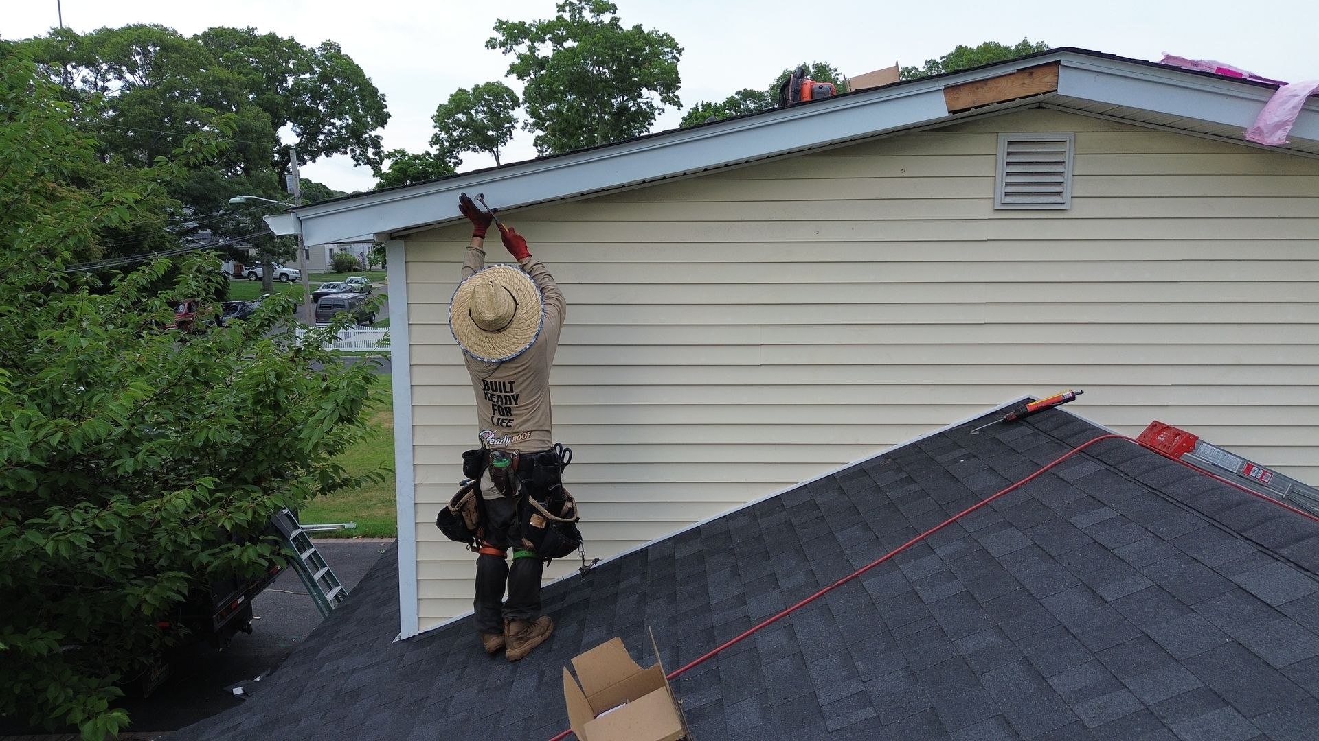 Worker in a straw hat on a roof beside a house, near trees and rooftop equipment.