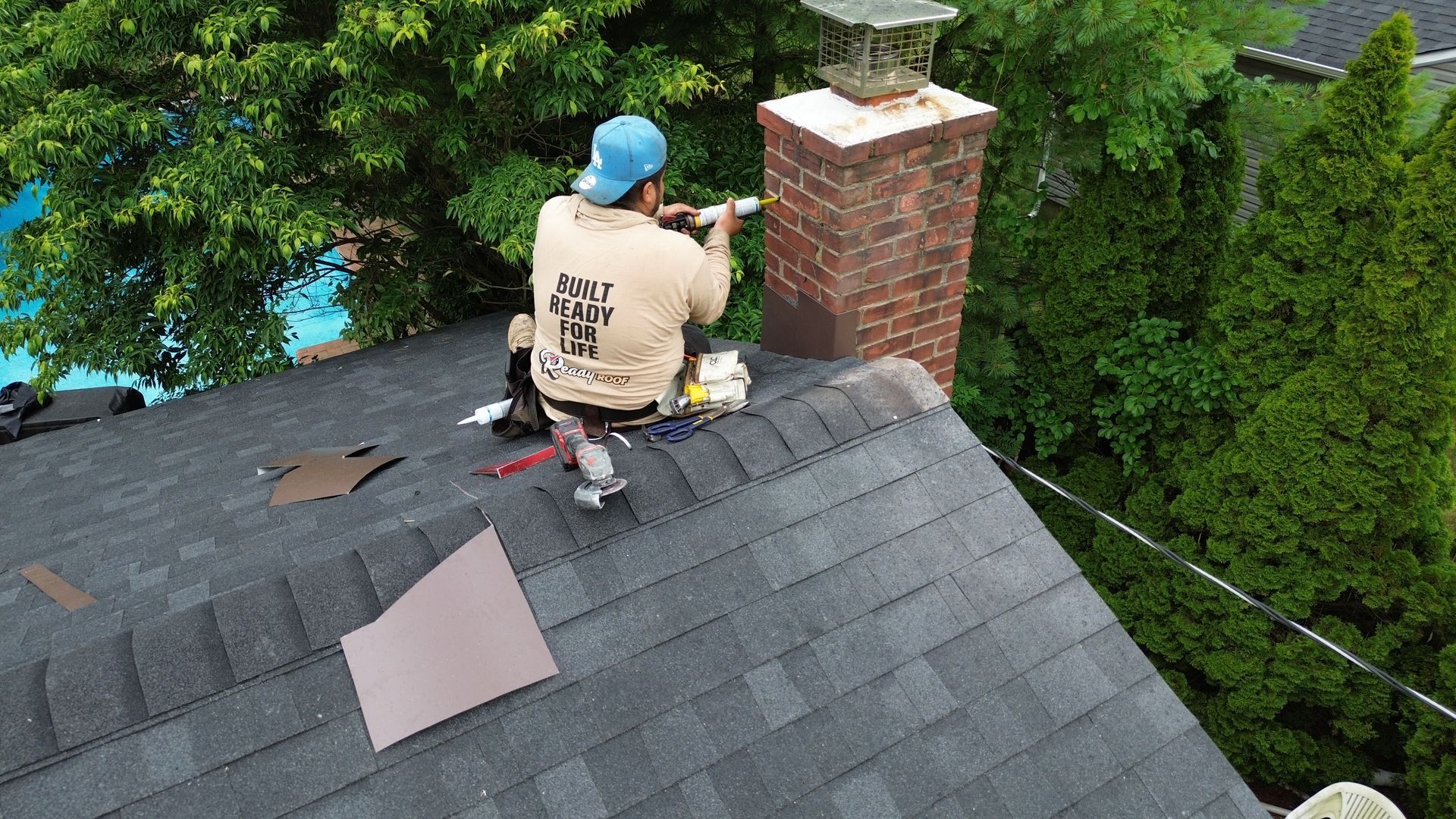 Worker repairing a shingled roof near a brick chimney, with tools on the roof and green trees nearby