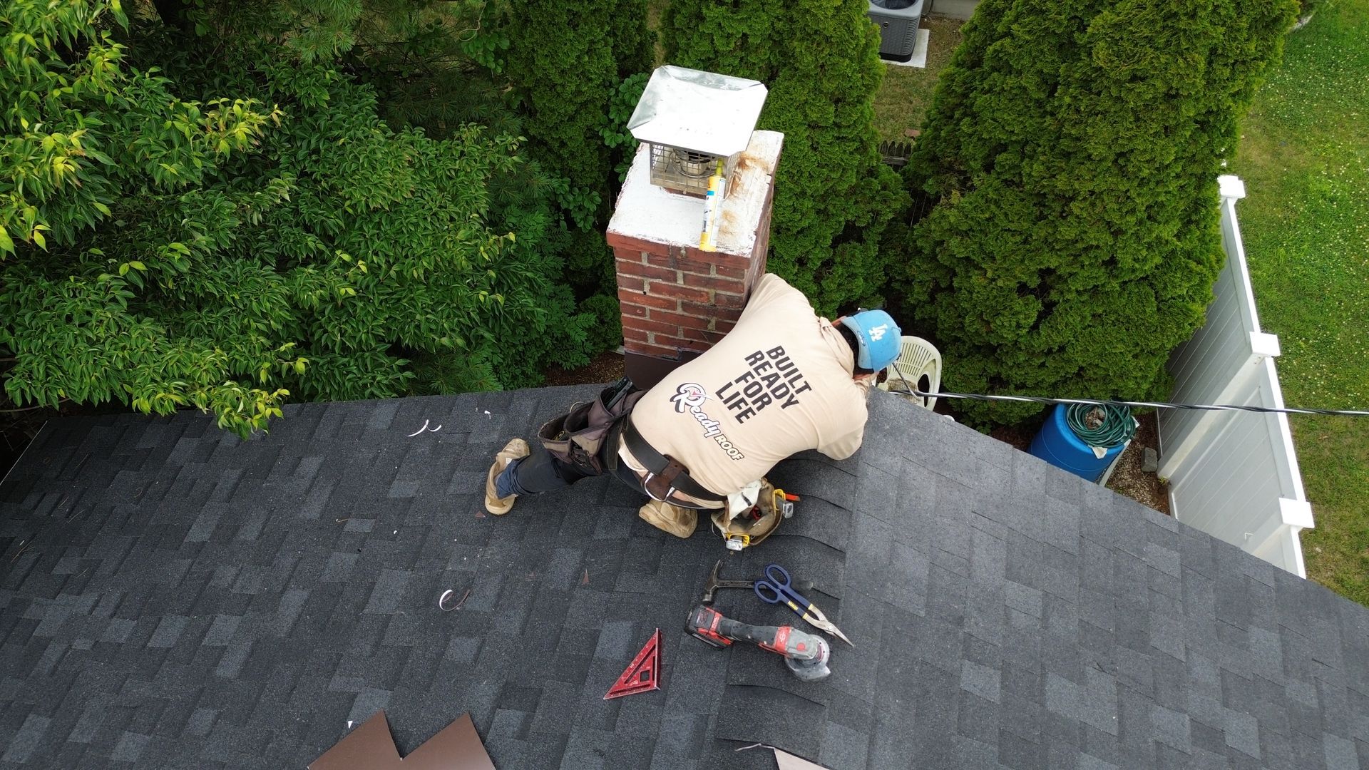 Roof worker on a shingled roof near a chimney, with tools and debris visible.