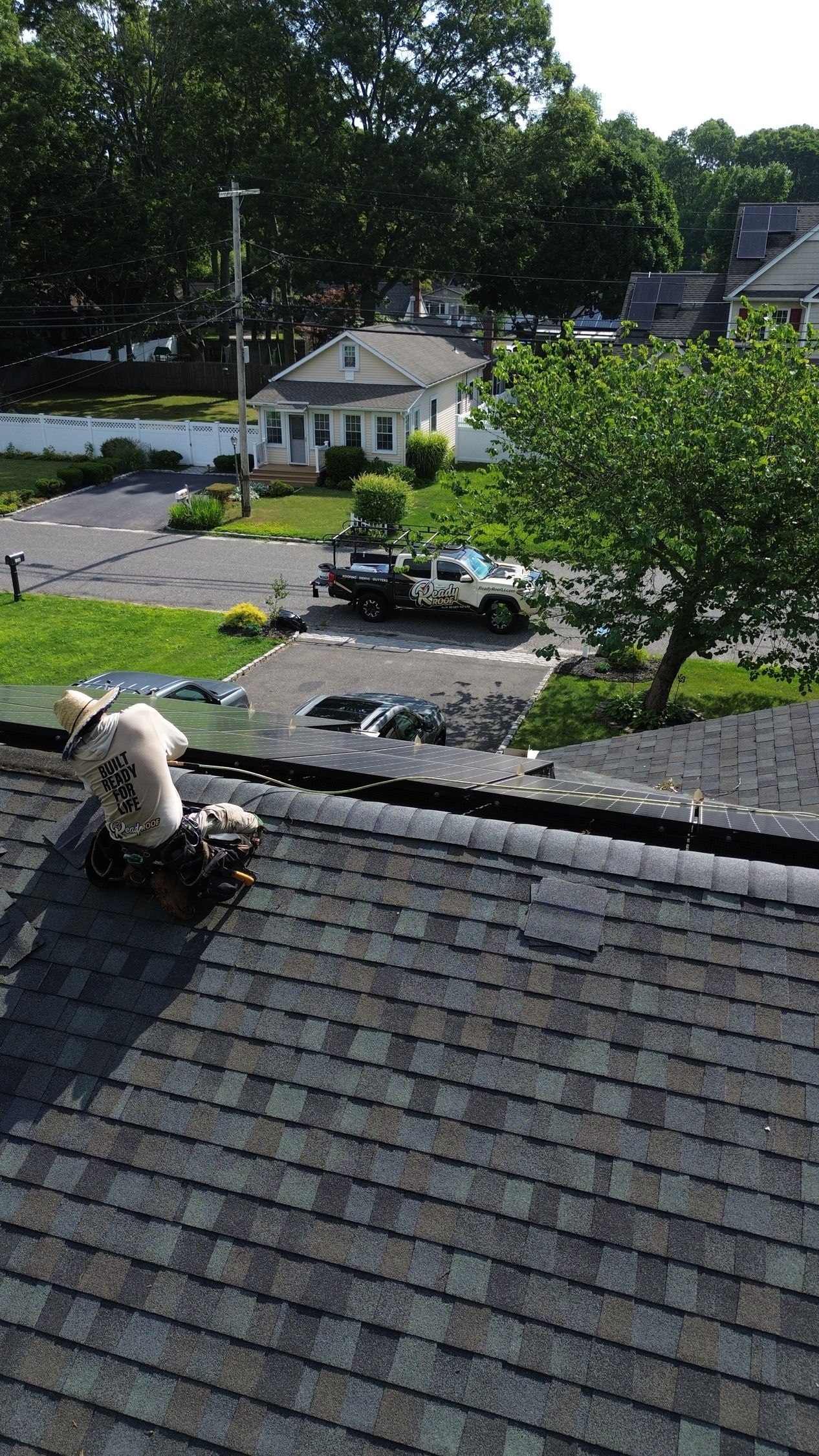 View from a shingled rooftop over a driveway, trees, and a small white house on a sunny day