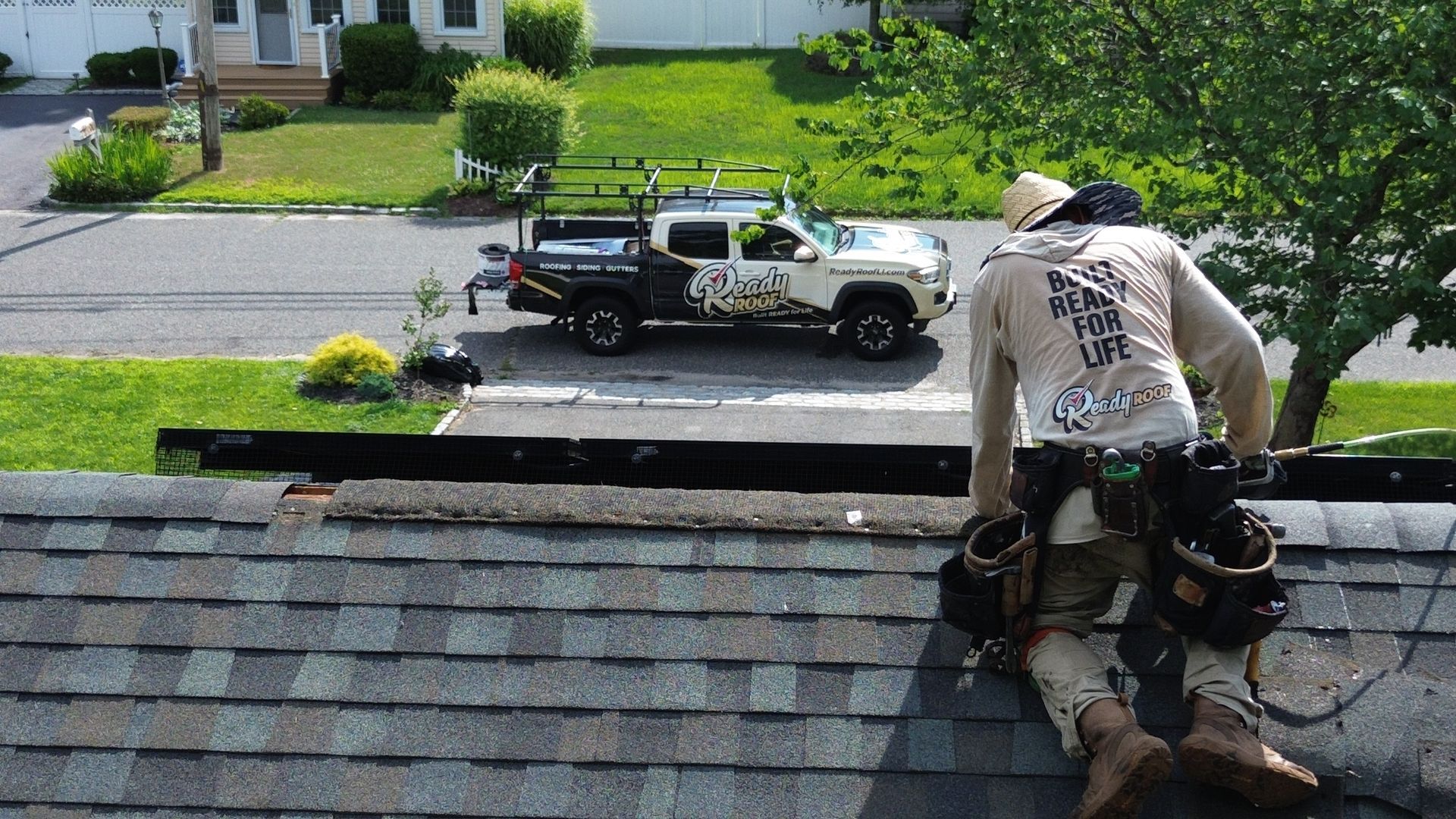 Worker on a shingled roof installing materials, with a truck and suburban street in the background