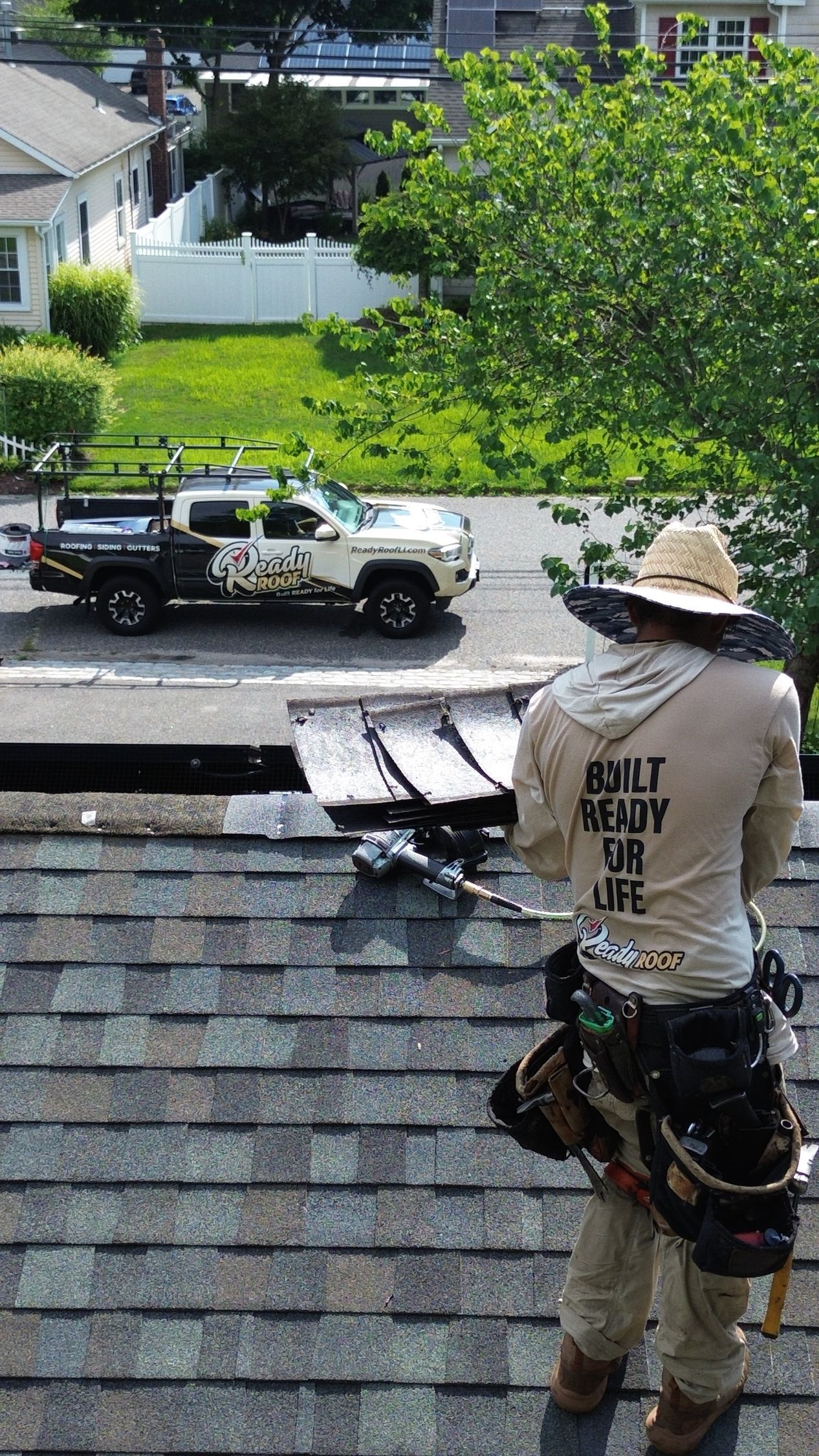 Worker on a roof with tools, facing a suburban street and parked truck.