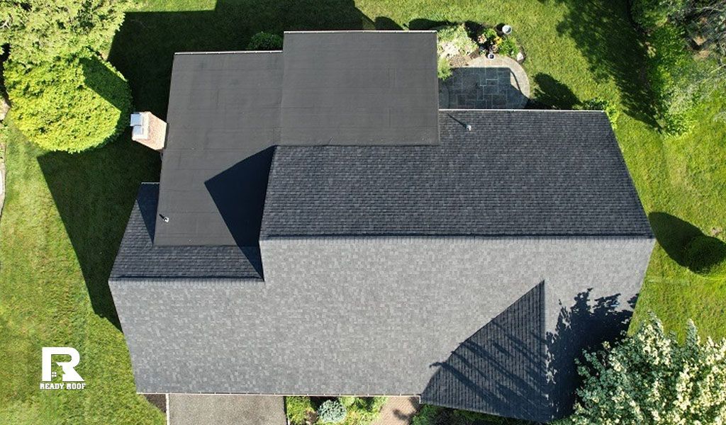 Aerial view of a house roof with gray shingles and a dark roof section, surrounded by green trees and lawn