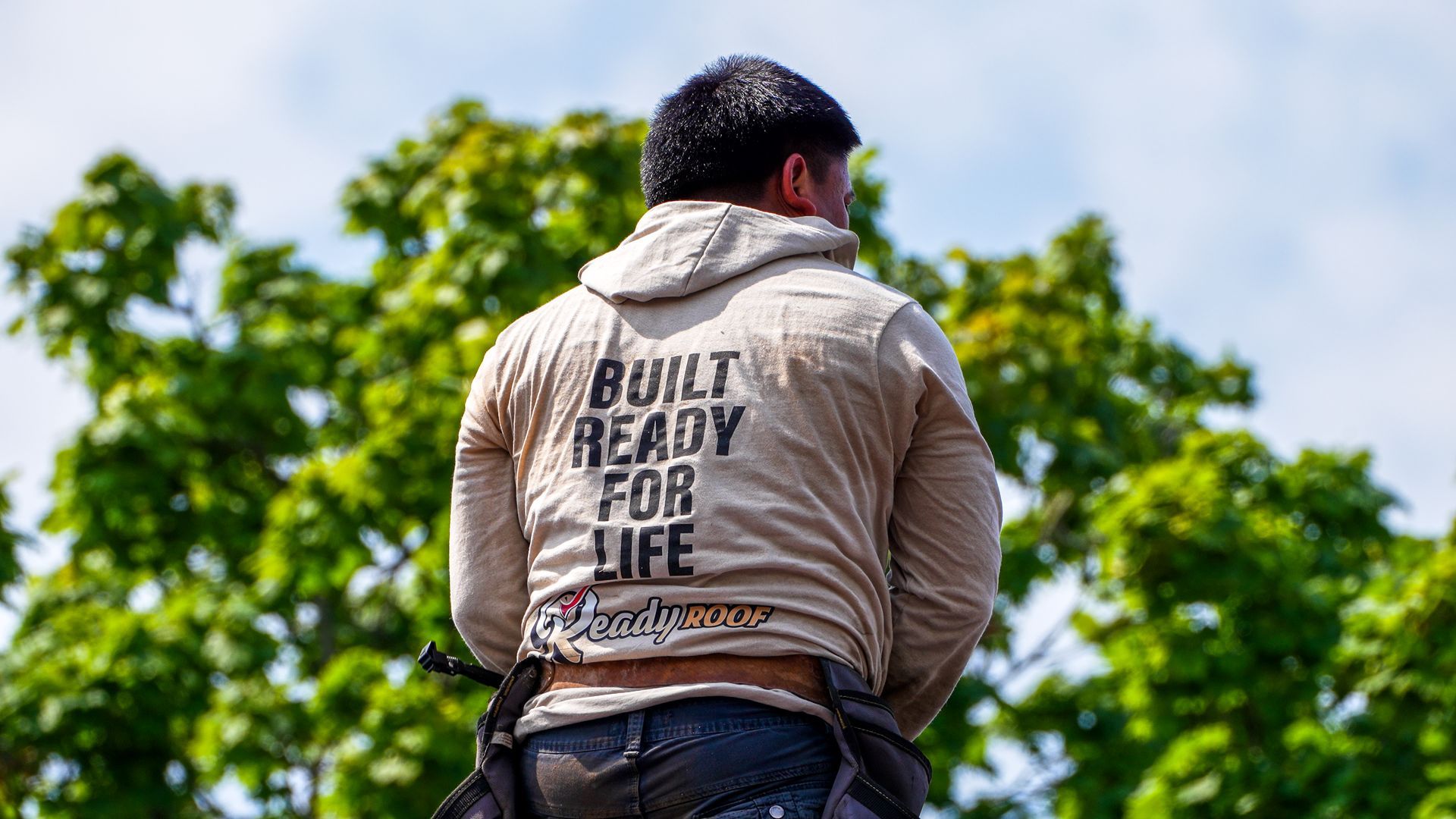 Person seen from behind wearing a tan hoodie with text, standing outdoors against green trees.