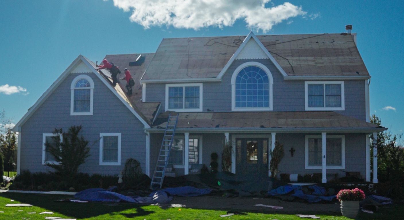 Blue two-story house with white trim and dormer windows under a bright sky, fronted by a porch and lawn.