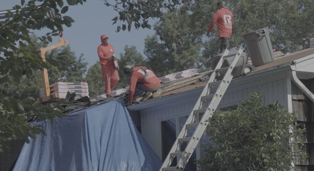Workers on a roof with ladders and a blue tarp, inspecting or repairing the house.