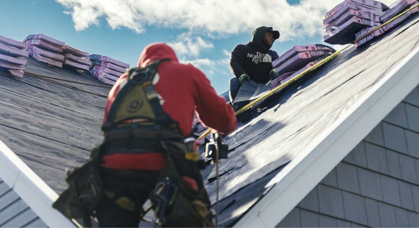 Two roofers in safety gear installing shingles on a steep roof, with stacked materials nearby.