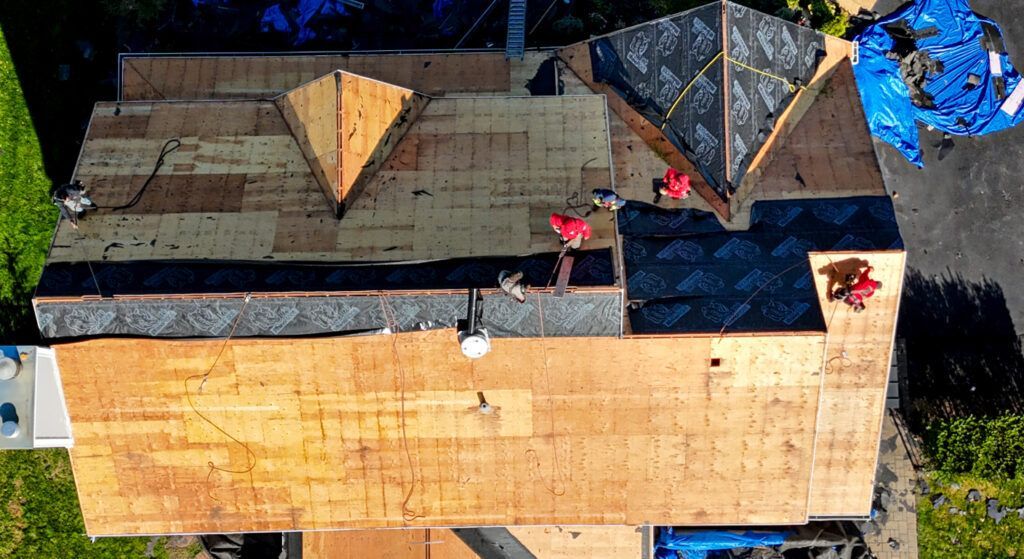 Aerial view of a house roof with dark shingles, plywood-covered sections, and blue tarps