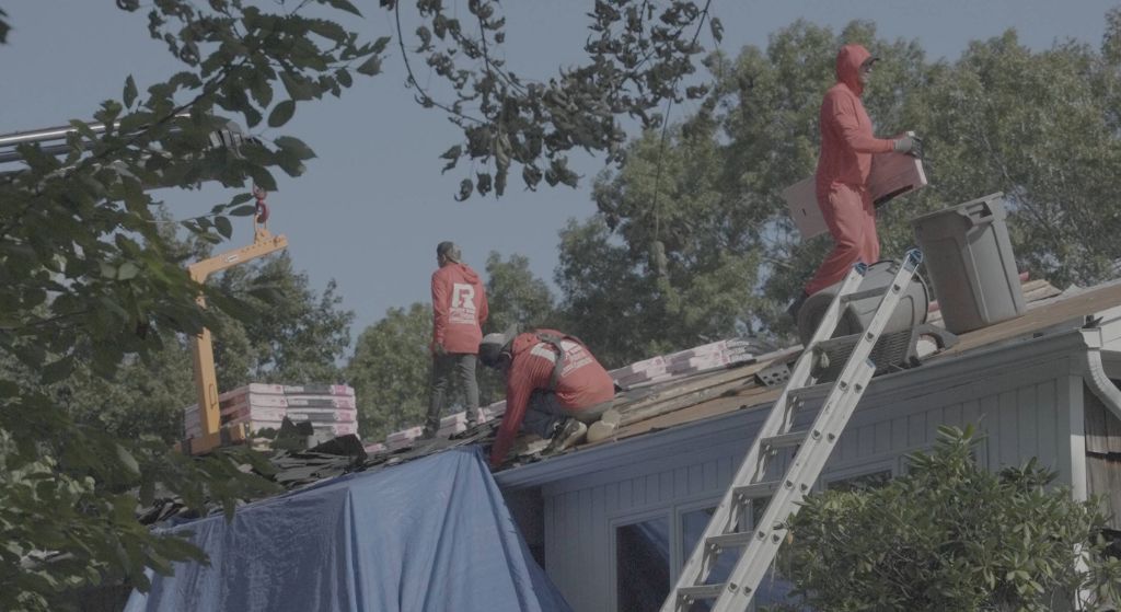 Roof workers in orange safety gear repair a damaged roof using a ladder and tarp.