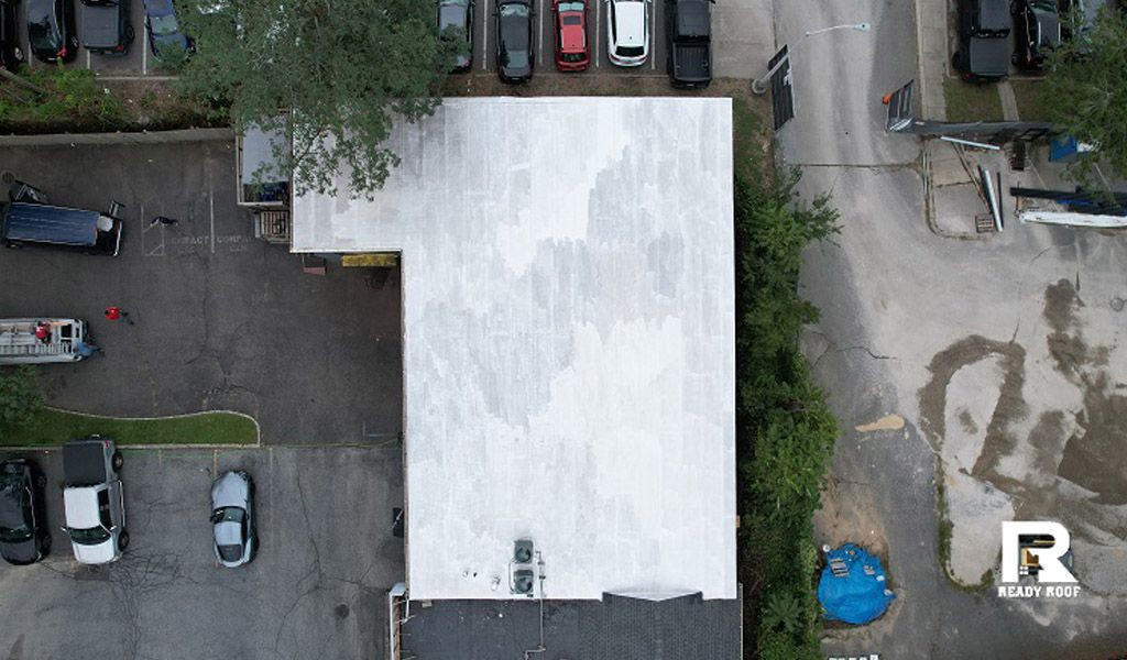 Aerial view of a white-roofed building surrounded by parked cars, trees, and a dirt lot