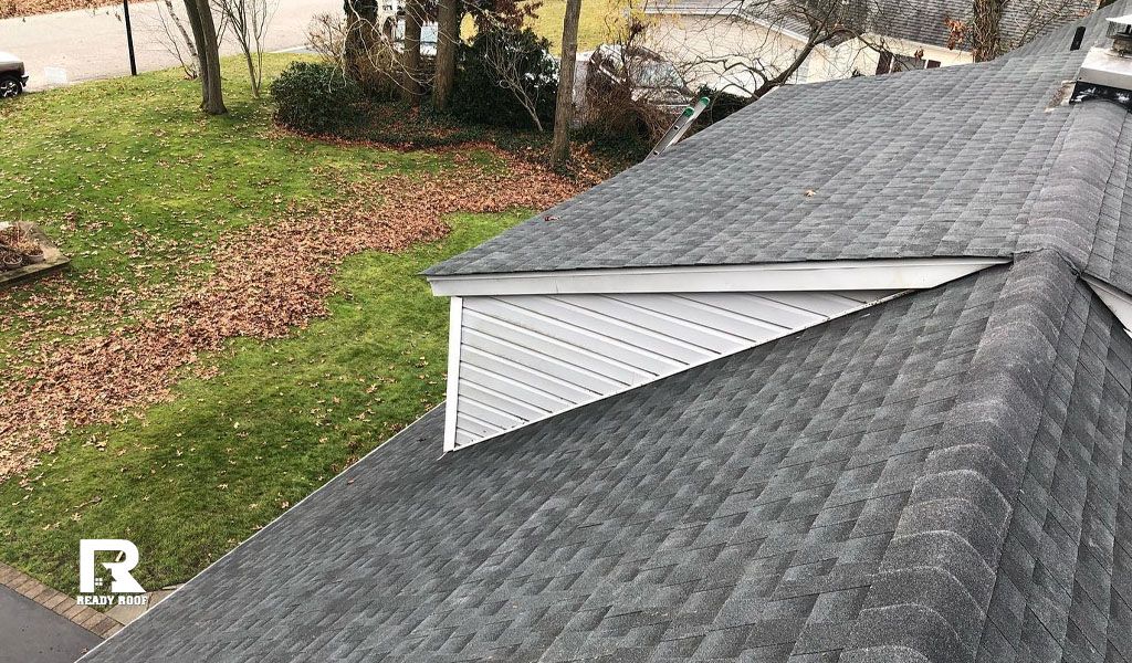 Roof with gray shingles and a small dormer on a grassy hillside near a driveway