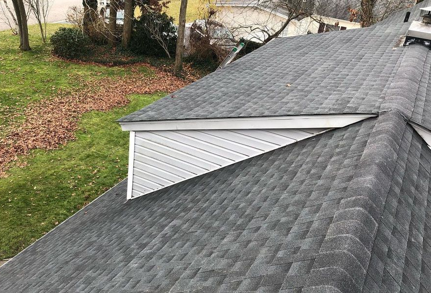Close-up of a gray shingled roof with a white dormer and sloped hillside in the background
