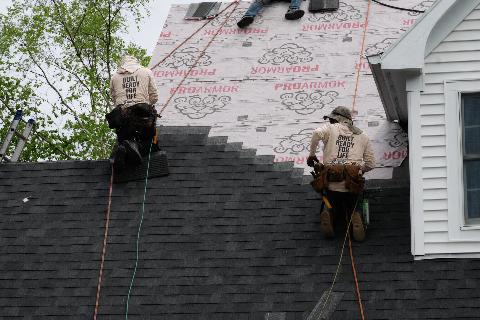 Two roofers installing black shingles on a house roof beside white siding and exposed underlayment
