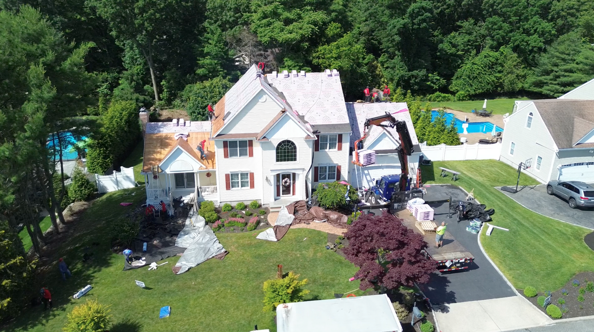 Aerial view of a large white house with a yard, driveway, patio, and swimming pool surrounded by trees.