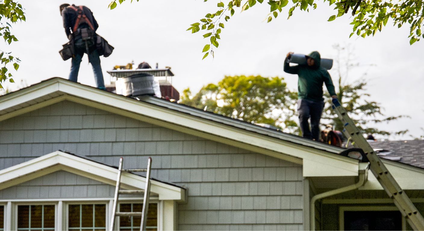Workers on a gray-roofed house repairing a chimney, with tools and materials on the roof.