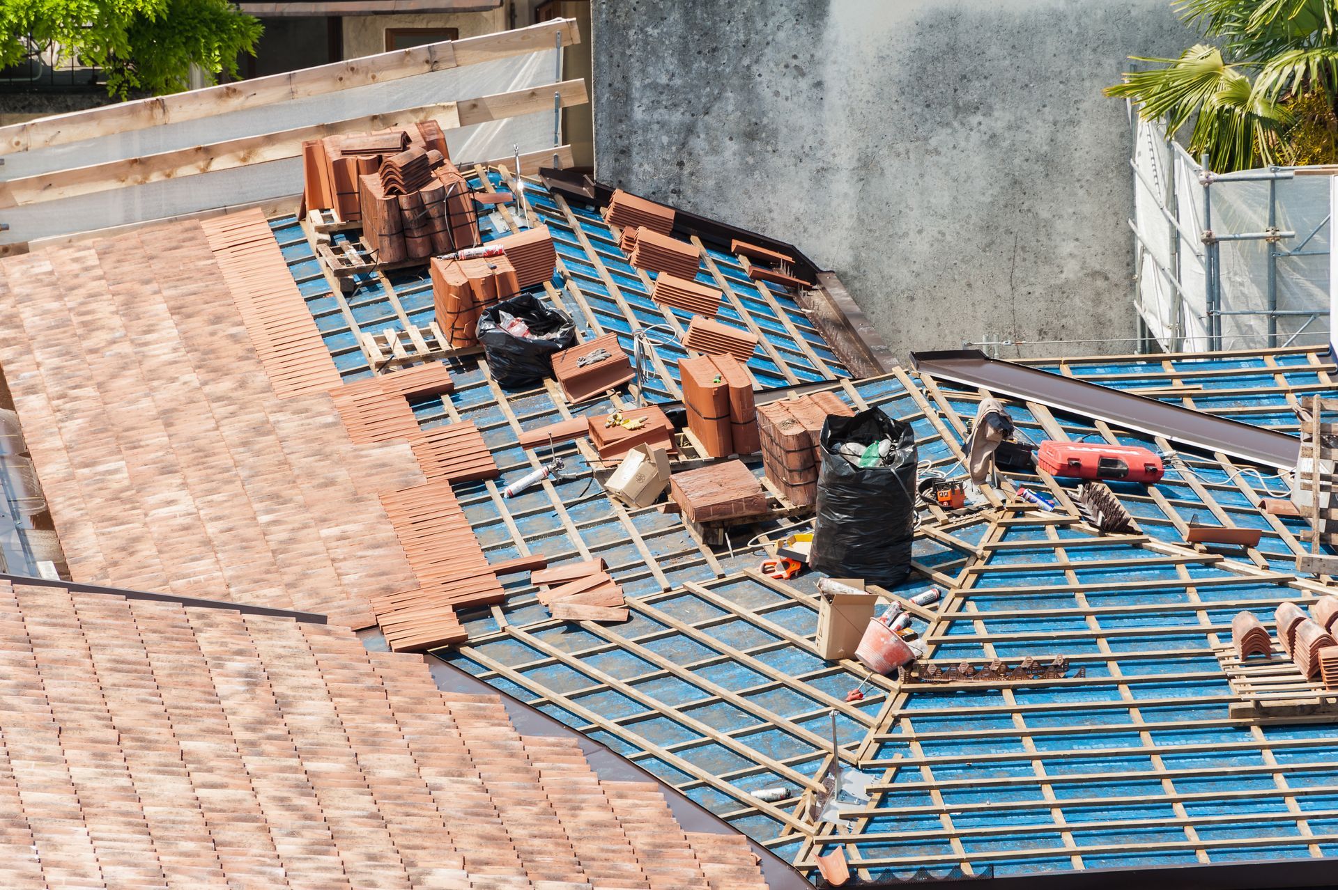Rooftop under construction with blue waterproofing mesh and stacks of red bricks beside a concrete wall.
