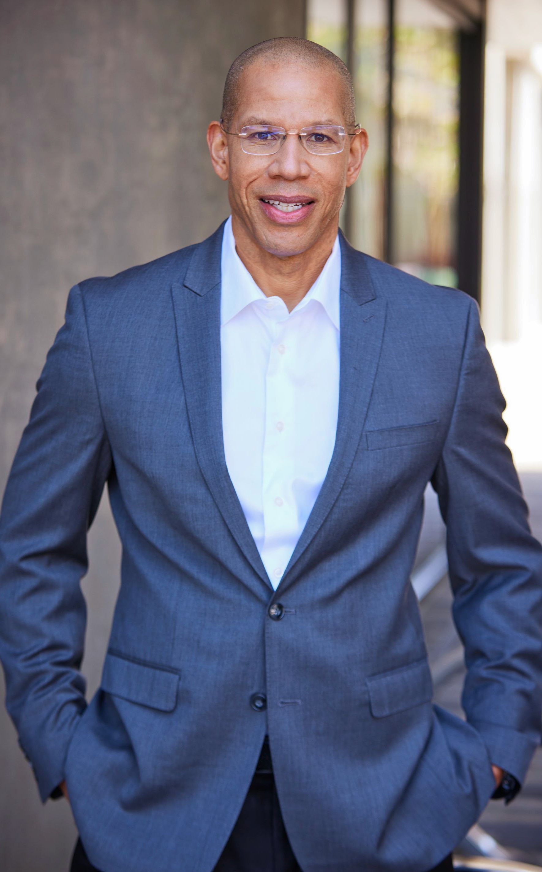 Man in gray suit smiles, hands in pockets, leaning against building, neutral background.