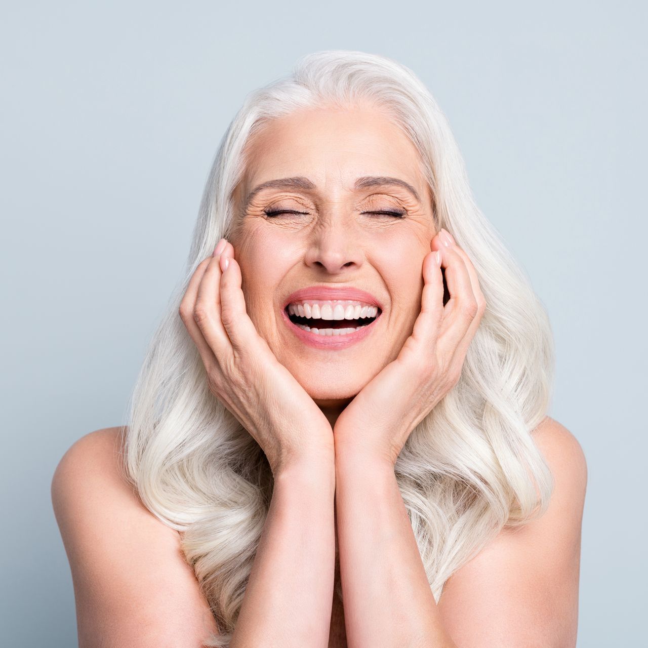 a woman with white hair is smiling with her eyes closed and her hands on her face .