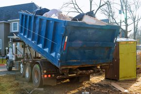 Blue dump truck unloading debris beside a yellow dumpster in a residential driveway