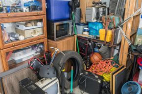 Cluttered garage storage area with shelves, tools, tires, wires, and assorted equipment.