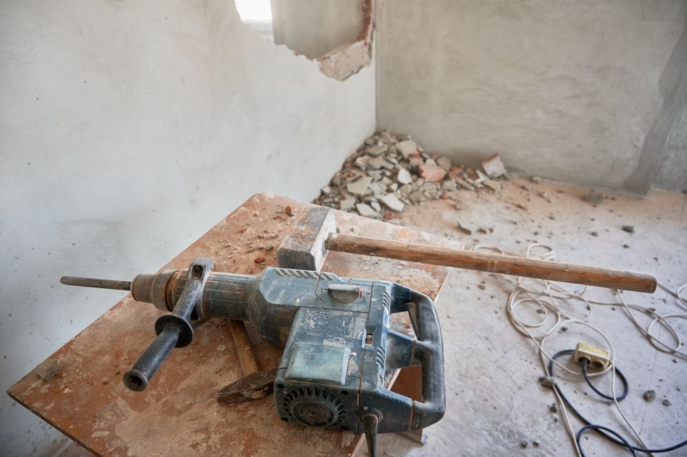 Jackhammer and tools on a dusty floor inside an unfinished room with rubble and a broken wall.