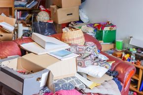 Cluttered living room with stacked boxes, papers, and assorted household items on a couch