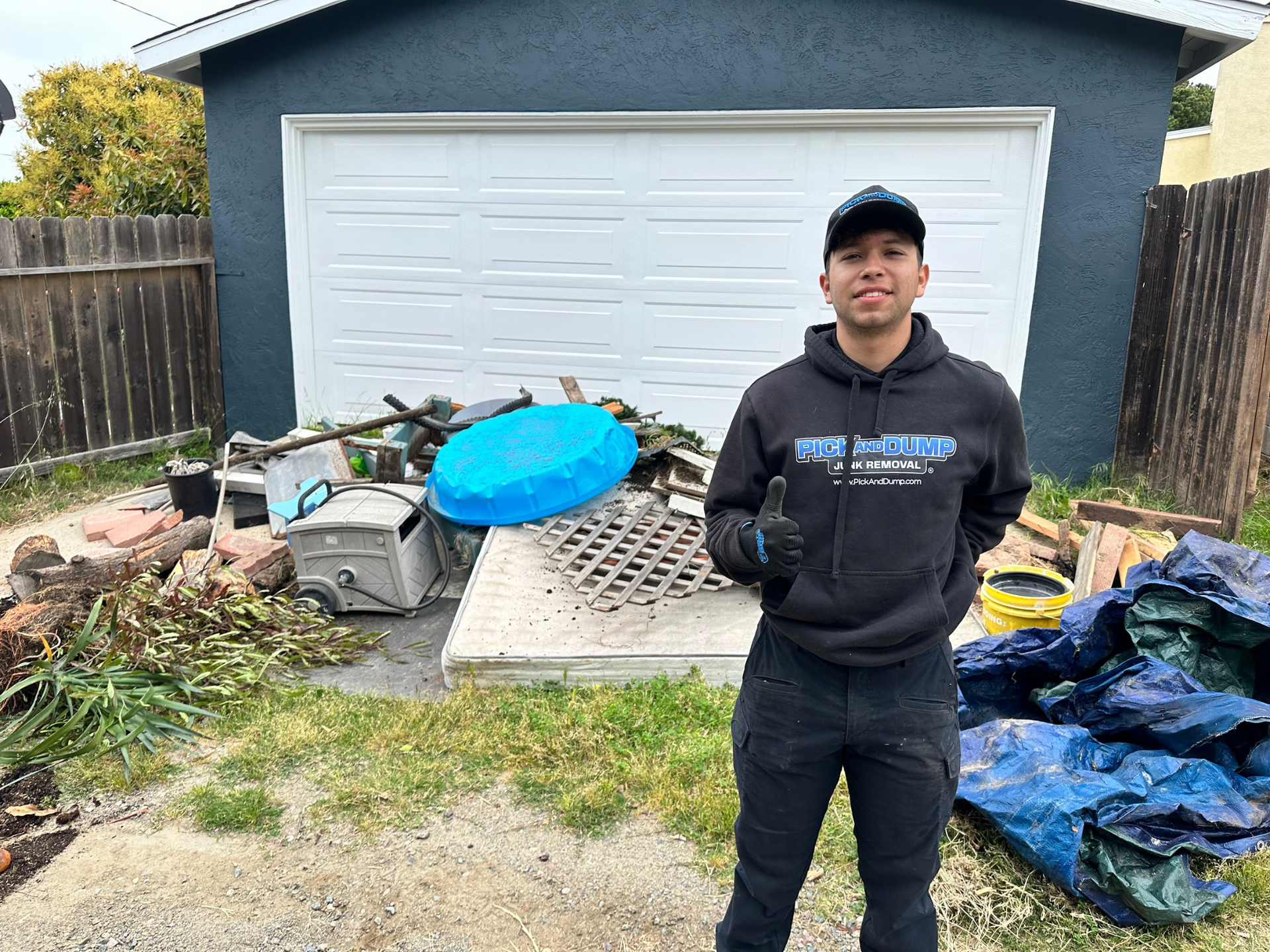 A man in a black hoodie is standing in front of a garage full of trash.